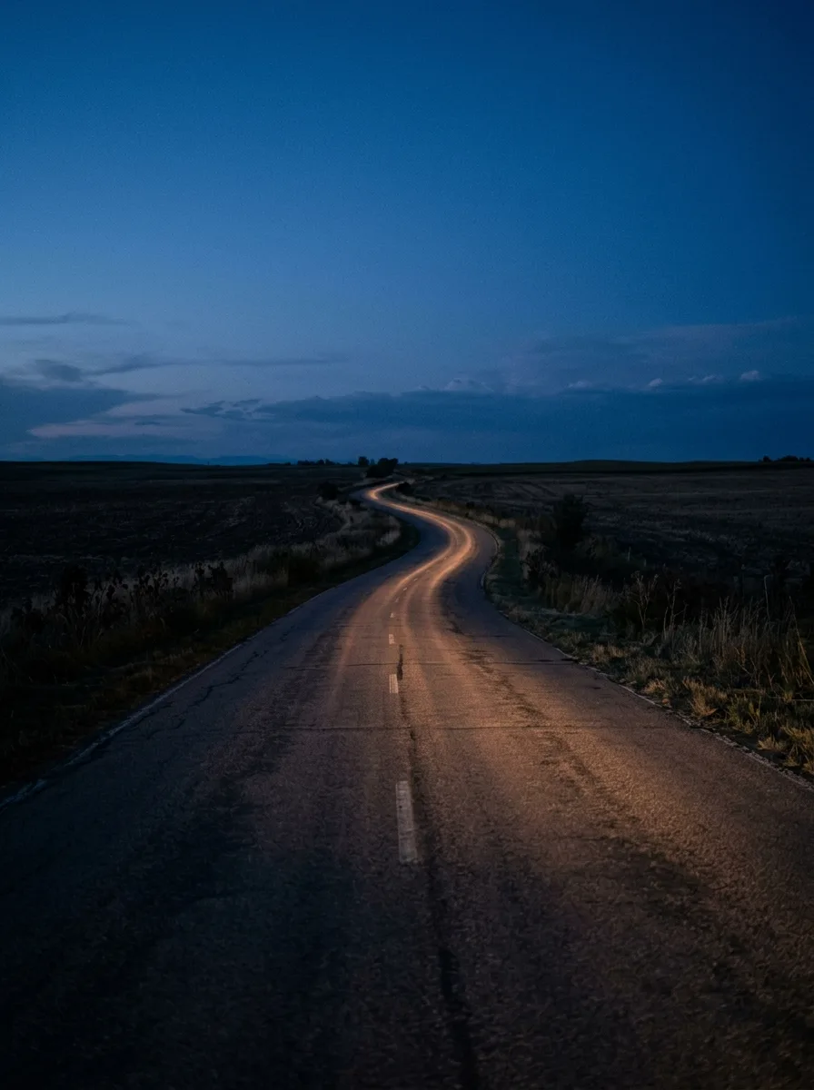 An empty two-lane road curving under a deep blue dusk sky, headlight glow on the asphalt